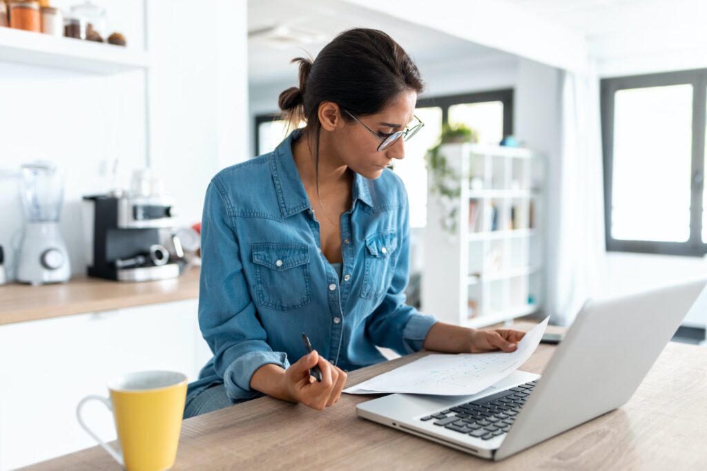 Pretty young woman reviewing paperwork and working on a laptop in the kitchen at home.